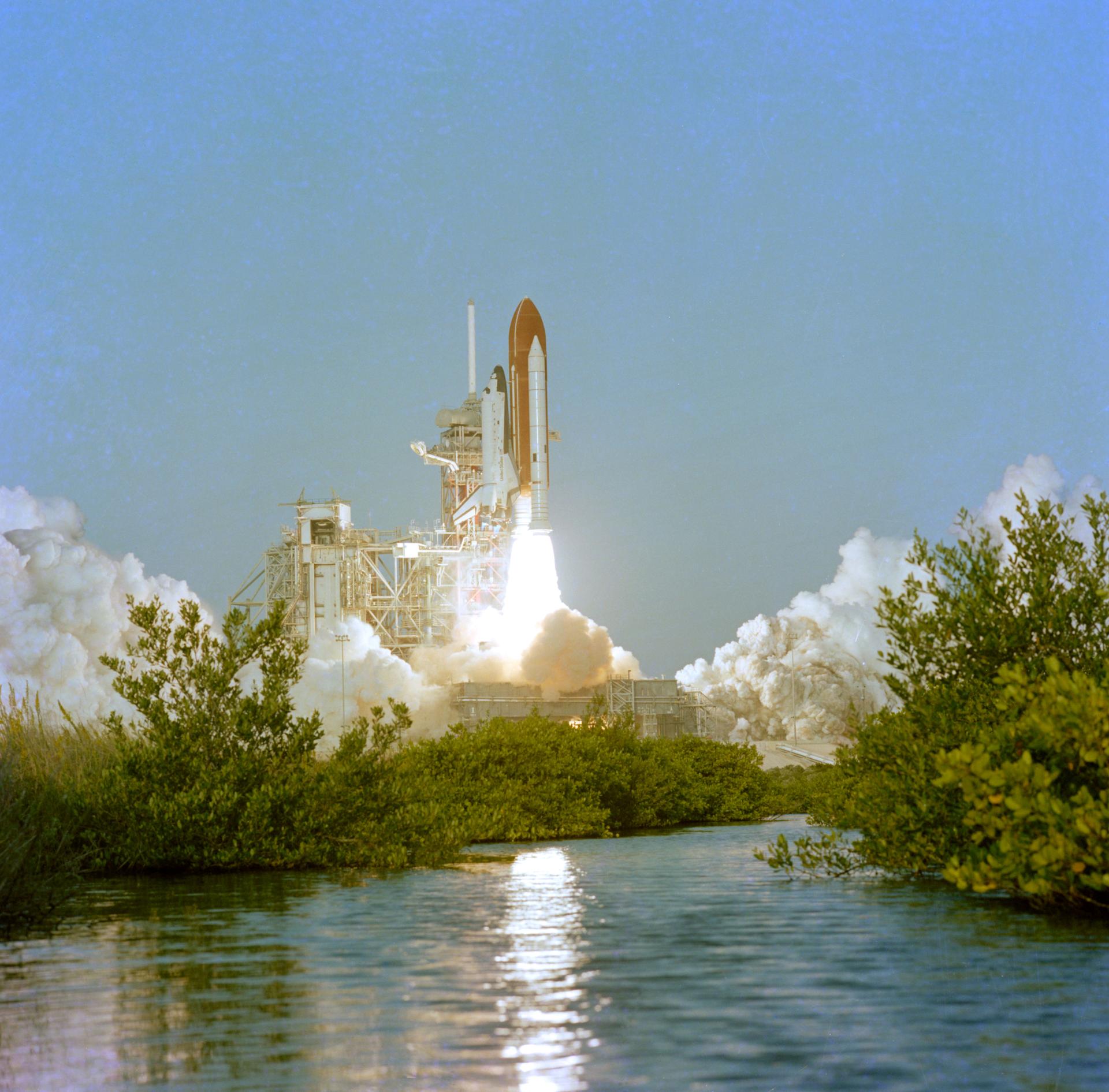 Space Shuttle Columbia launches into a cloudless blue sky from Kennedy Space Center. Water and vegetation are visible in the foreground.