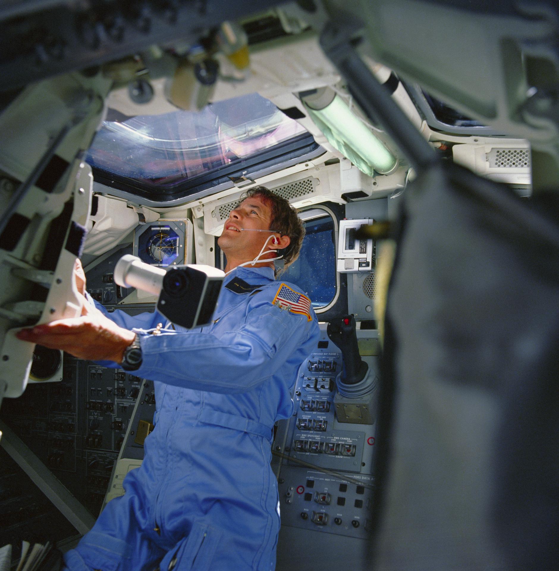 Dr. Joseph Allen is seen looking out the Space Shuttle window with a spot meter floating next to him