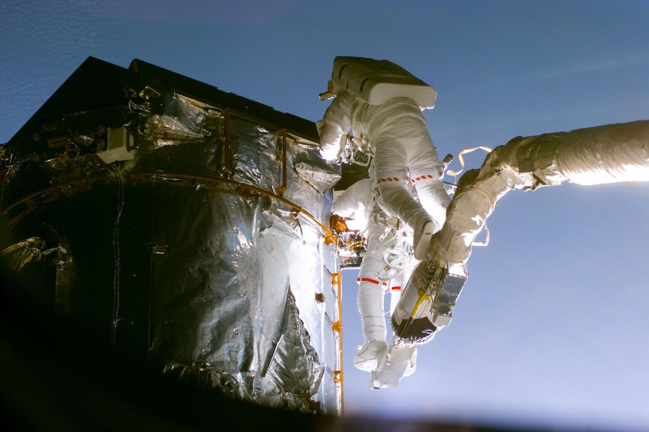 S82-E-5652 (17 Feb. 1997) --- Astronaut Gregory J. Harbaugh (solid stripe on EMU) uses Remote Manipulator System (RMS) as a cherry-picker device to service Hubble Space Telescope (HST). In cooperation with astronaut Joseph R. Tanner, nearby, the mission specialist was in the process of replacing the HST's Magnetic Sensing System (MSS) protective caps with new, permanent covers.  This view was taken with an Electronic Still Camera (ESC).