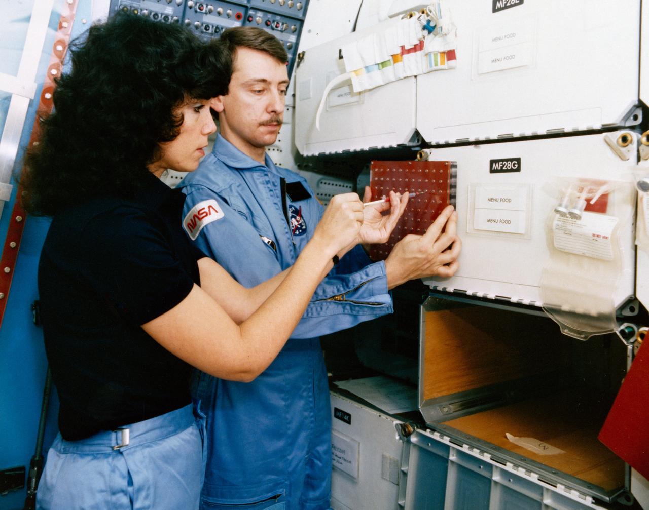S84-35757 (May 1984) --- Astronaut Judith A. Resnik, 41-D mission specialist, and Charles Walker, payload specialist for that June 1984 flight, prepare for some scheduled intravehicular activity involving the continuous flow electrophoresis systems (CFES) experiment.  CFES will join the six-member crew aboard the Earth-orbiting Discovery for a seven day mission.  The two share in preparing a sample to be processed by the CFES.  In the background are stowage lockers and a CFES trainer-- part of the Shuttle one-g trainer at NASA's Johnson Space Center (JSC).  Walker, an engineer at McDonnell Douglas Astronautics Co. in St. Louis, Missouri, will be the first Shuttle payload specialist to represent a project designed for commercial purposes.  As payload specialist, his job will be to run the materials electrophoresis-operations-in-space project.  The project is aimed at separating large quantities of biological materials in space for ultimate use in new pharmaceuticals.  The photo was taken by a McDonnell Douglas photographer.