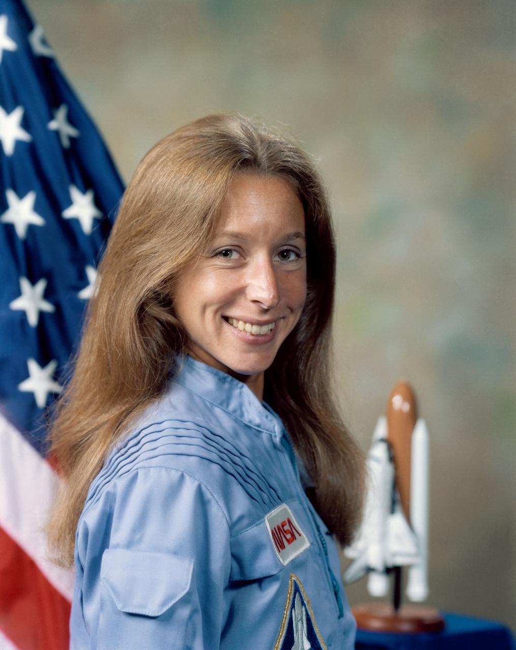 Portrait photograph, Astronaut Marsha S. Ivins, dressed in Blue Flight Suit, with Flag (frame left), and Space Shuttle Model (frame right).                         JSC, HOUSTON, TX