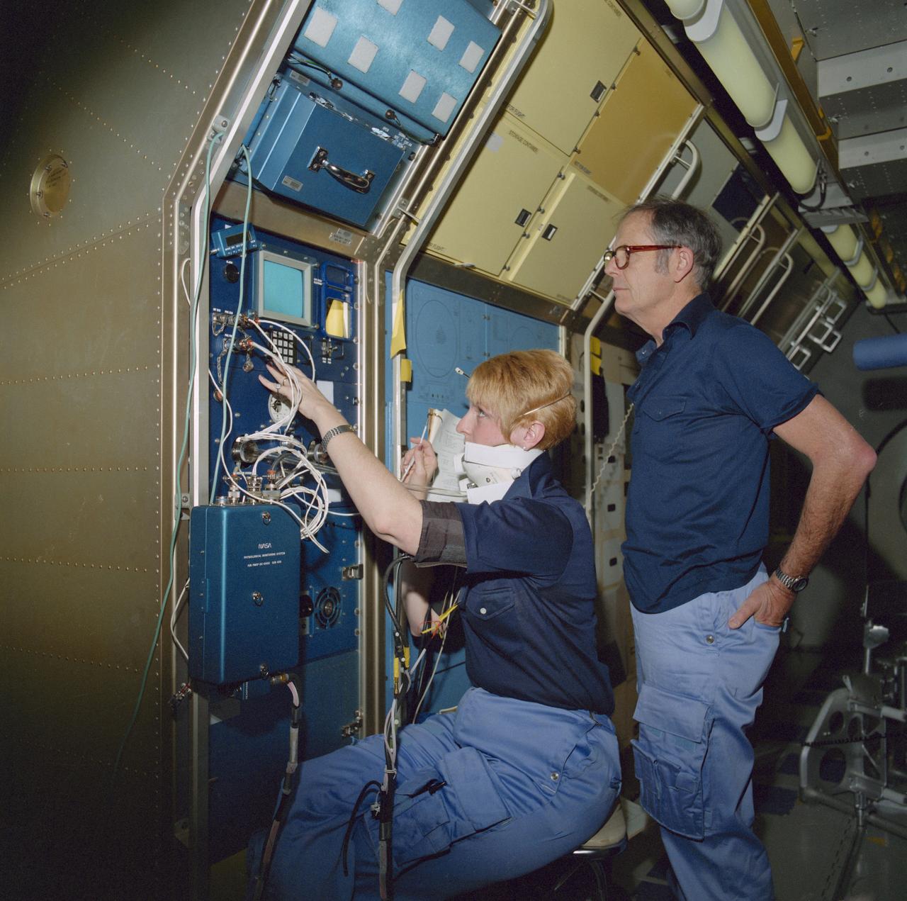 S85-26571 (Feb 1985) --- Wearing a special collar, Millie Hughes-Fulford, payload specialist, practices medical test operations scheduled for the Spacelab Life Sciences (SLS-1) mission. Robert Ward Phillips, backup payload specialist, looks on. The collar, called the baroflex neck pressure chamber, is designed to stimulate the bioceptors in the carotid artery, one of the two main arteries that supply blood to the head.
