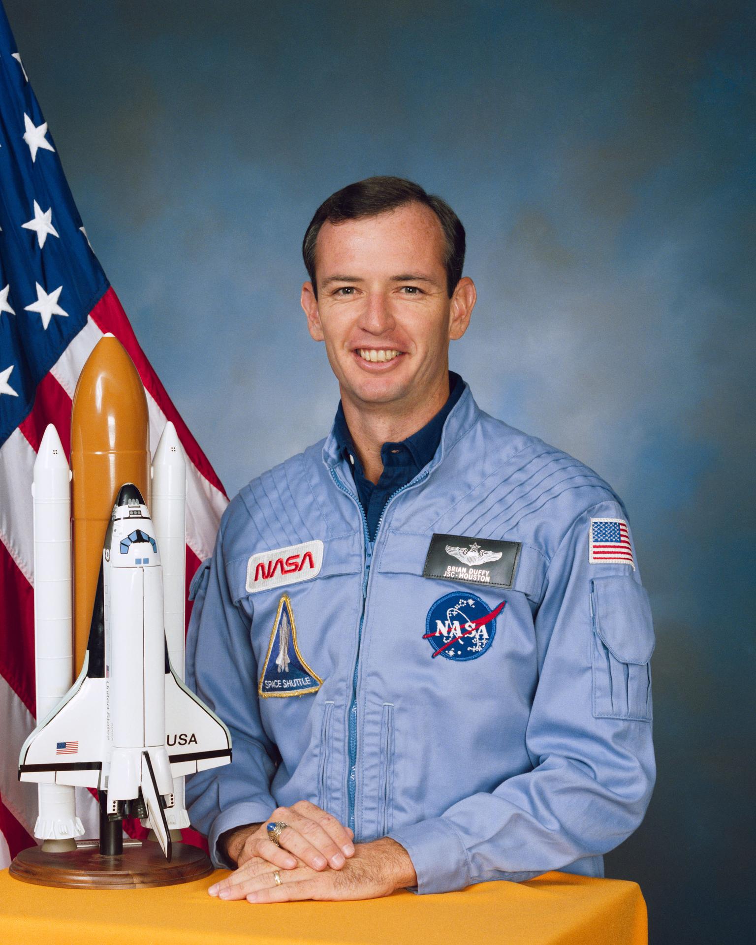 A man smiles, wearing an astronaut flight suit. He is sitting in front of the American flag and beside a model rocket.