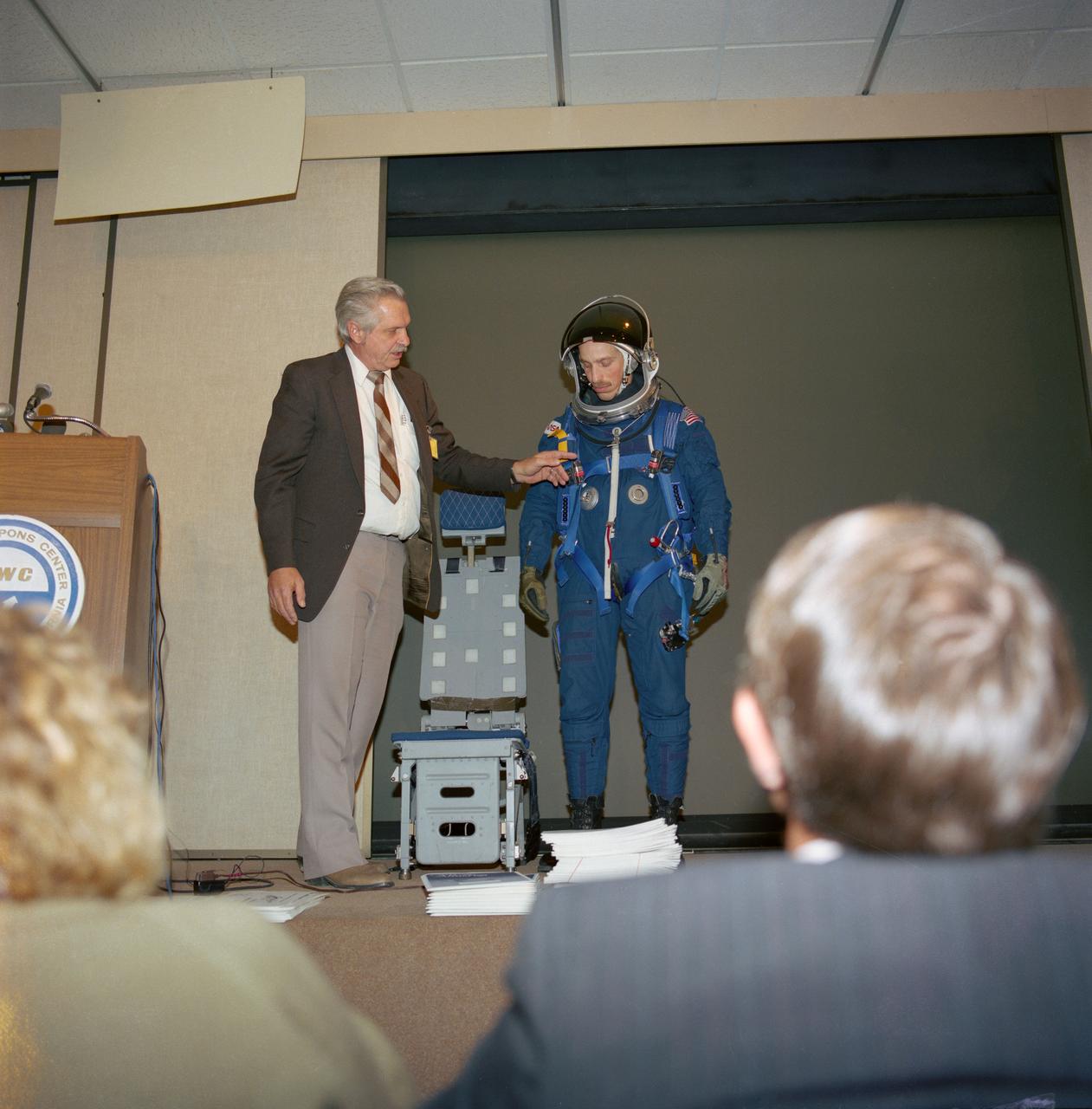 S88-25408 (8 Dec 1987) --- James O. Schlosser (left), JSC crew systems branch employee responsible for crew equipment development, gives a briefing on the crew equipment baselined for STs-26 as astronaut James P. Bagian models the new gear.  Included in the package are a partial pressure suit, harness, parachute, life raft and survival gear.  The deomonstration took place at the Naval Weapons Center in China Lake, CA.