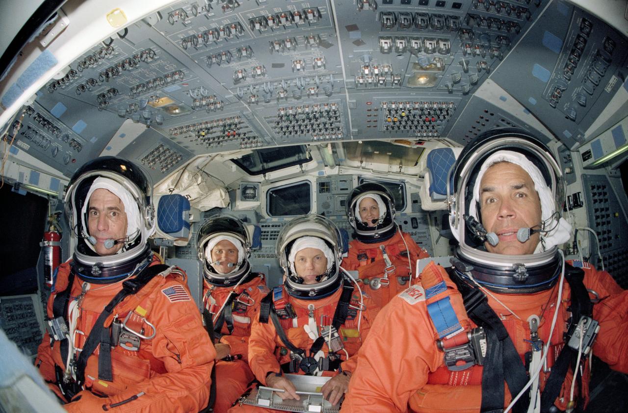 STS-33 Discovery, Orbiter Vehicle (OV) 103, crewmembers, wearing orange launch and entry suits (LESs) and launch and entry helmets (LEHs), are seated in their launch and entry positions on crew compartment trainer (CCT) flight deck during a training exercise in JSC Mockup and Integration Laboratory (MAIL) Bldg 9A. Commander Frederick D. Gregory (far right) is stationed at forward flight deck commanders controls, Pilot John E. Blaha (far left) at the pilots controls and on aft flight deck are mission specialists Manley L. Carter, Jr (left), MS F. Story Musgrave (center, holding clipboard), and MS Kathryn C. Thornton (standing). Overhead forward control panels are visible above the astronauts and aft flight deck onorbit station control panels and windows are visible in the background. Thornton is on the flight deck for this photo but during launch and entry will be seated on the middeck.