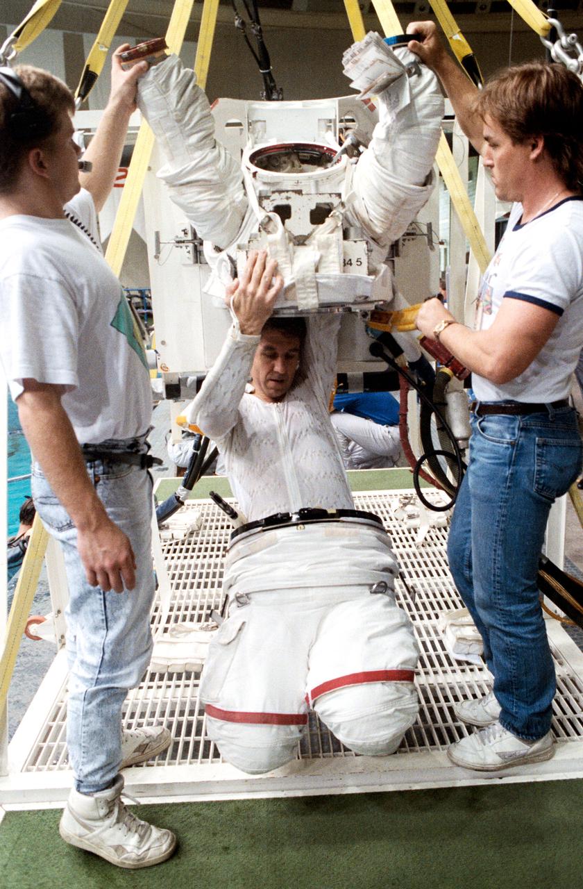STS-38 Mission Specialist (MS) Robert C. Springer dons extravehicular mobility unit (EMU) upper torso with technicians' assistance in JSC's Weightless Environment Training Facility (WETF) Bldg 29. Positioned on the WETF platform at pool side, Springer is preparing for an underwater extravehicular activity (EVA) simulation. During the training session, Springer will rehearse contingency EVA procedures for the STS-38 mission aboard Atlantis, Orbiter Vehicle (OV) 104.