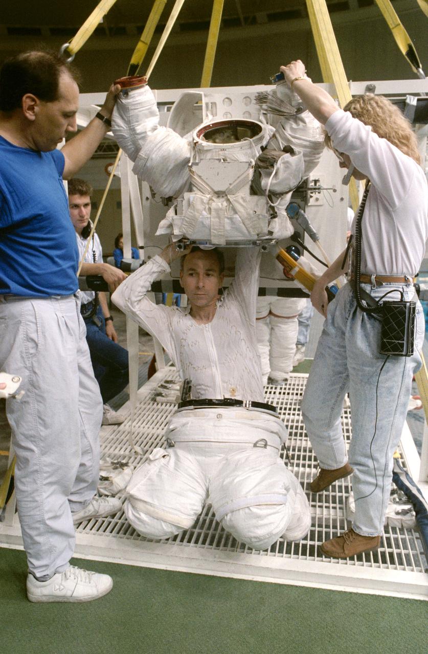 STS-38 Mission Specialist (MS) Carl J. Meade, wearing liquid cooling and ventilation garment (LCVG) and extravehicular mobility unit (EMU) lower torso, crouches under EMU upper torso. Technicians extend the EMU sleeves as Meade reaches into upper torso during suit donning in JSC's Weightless Environment Training Facility (WETF) Bldg 29. Positioned on the WETF platform at pool side, Meade is preparing for an underwater extravehicular activity (EVA) simulation. During the training exercise, Meade will rehearse contingency EVA procedures for the STS-38 mission aboard Atlantis, Orbiter Vehicle (OV) 104.