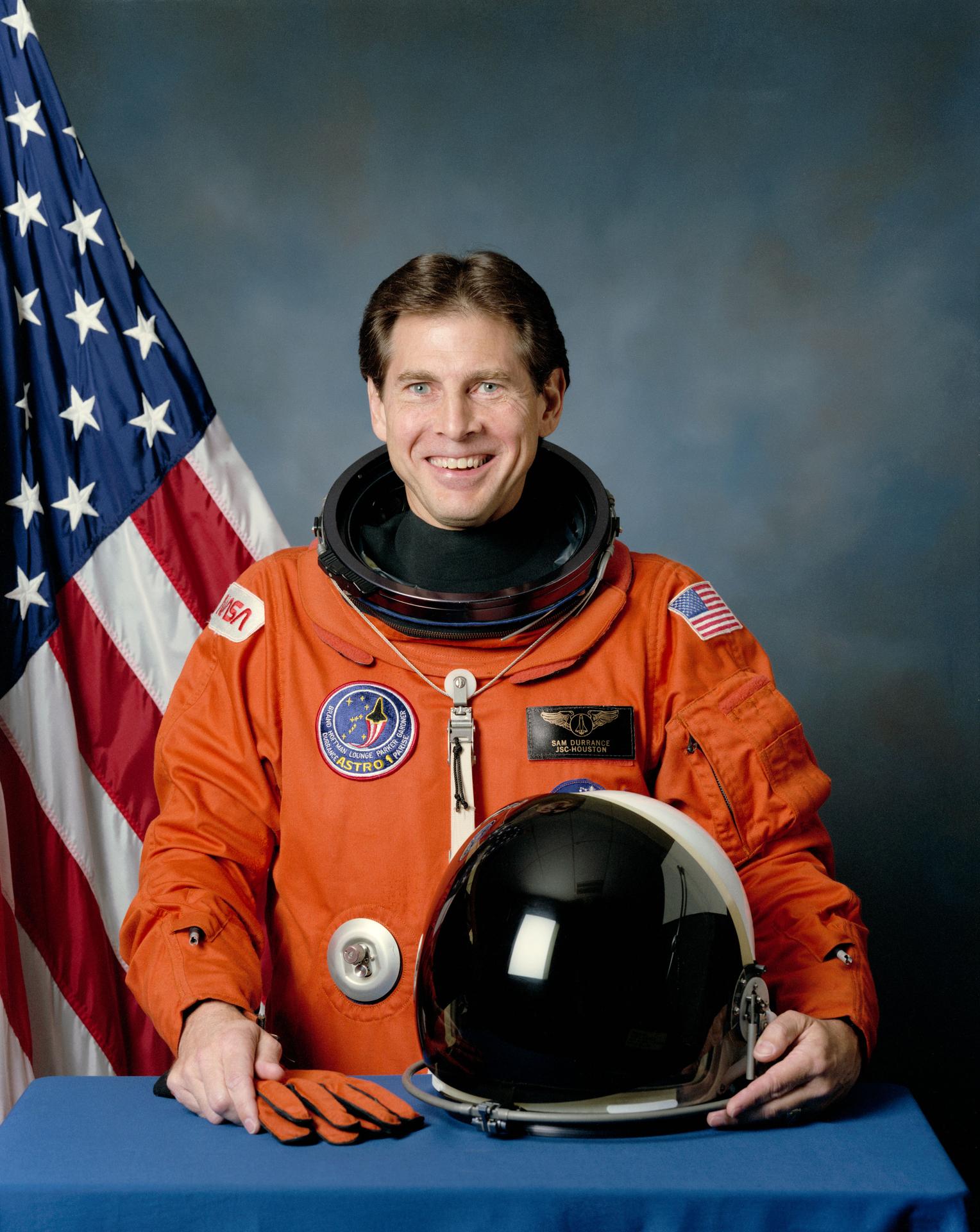 A man smiles, wearing a spacesuit, and stands in front of the American flag. He has his helmet off and sitting on table before him.