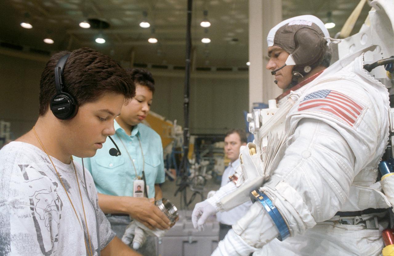 S92-42681 (28 July 1992) --- STS-53 Discovery, Orbiter Vehicle (OV) 103, Mission Specialist (MS) Michael R.U. Clifford, wearing extravehicular mobility unit (EMU) and communications  carrier assembly (CCA), dons gloves with assistance from two technicians.   Clifford is preparing for an underwater contingency extravehicular activity  (EVA) simulation in JSC's Weightless Environment Training Facility (WETF)  Bldg 29 pool.