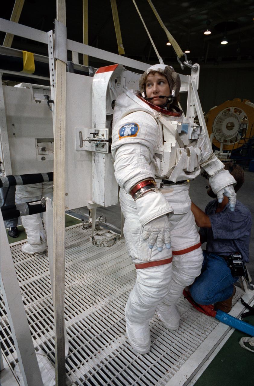 S92-42753 (31 July 1992) --- Astronaut Susan J. Helms, mission specialist assigned to fly aboard the Space Shuttle Endeavour for the STS-54 mission, gets assistance to complete the donning of her spacesuit. Though not assigned to the scheduled extravehicular activity (EVA), Helms is trained in the weightless environment training facility (WET-F). She will aid astronauts Gregory J. Harbaugh and Mario Runco Jr. in their planned EVA, scheduled for January of next year, and serve a backup role. Wearing this high fidelity training version of the extravehicular mobility unit (EMU), Helms was later lowered into the 25-ft. deep WET-F pool. The pressurized suit is weighted so as to allow Helms to achieve neutral buoyancy and simulate the various chores of the spacewalk.
