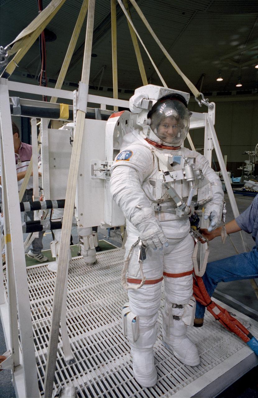 S92-42754 (31 July 1992) --- Astronaut Susan J. Helms, mission specialist assigned to fly aboard the Space Shuttle Endeavour for the STS-54 mission, gets assistance to complete the donning of her spacesuit. Though not assigned to the scheduled extravehicular activity (EVA), Helms is trained in the weightless environment training facility (WET-F). She will aid astronauts Gregory J. Harbaugh and Mario Runco Jr. in their planned EVA, scheduled for January of next year, and serve a backup role. Wearing this high fidelity training version of the extravehicular mobility unit (EMU), Helms was later lowered into the 25-ft. deep WET-F pool. The pressurized suit is weighted so as to allow Helms to achieve neutral buoyancy and simulate the various chores of the spacewalk.