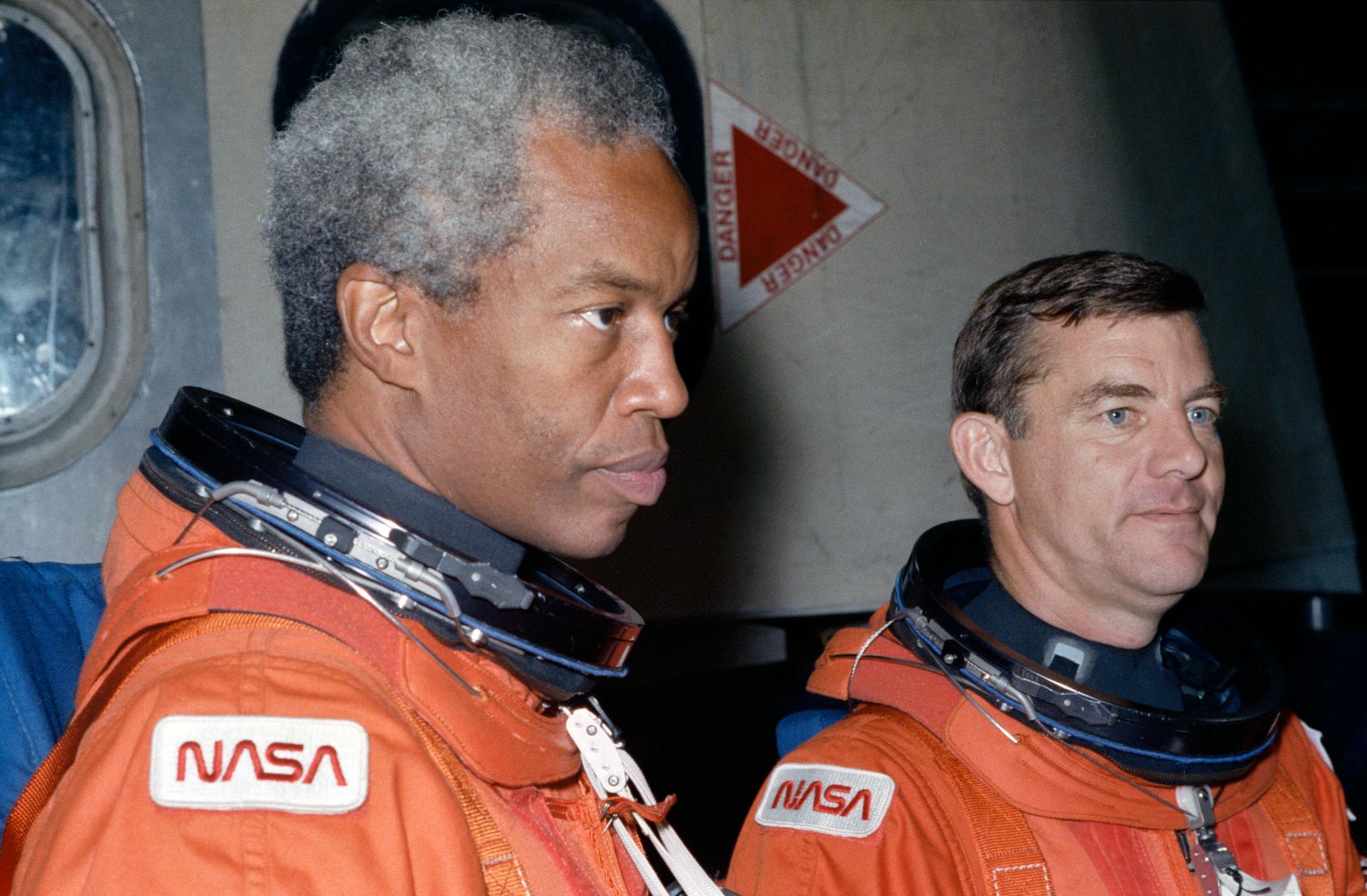 Astronaut Guion Bluford wearing an orange flight suit listens to instructions.