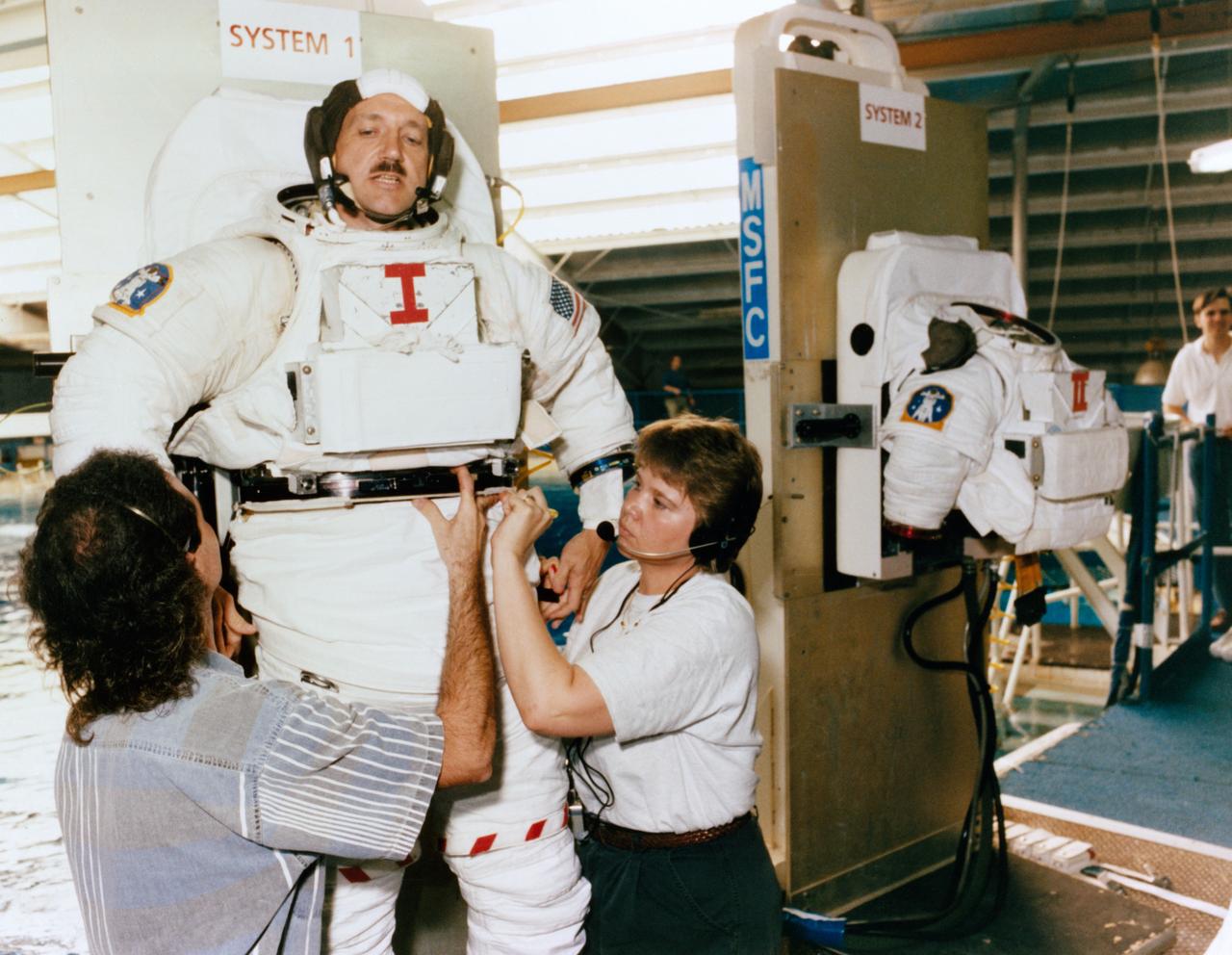 Astronaut Thomas D. Akers gets assistance in donning a training version of the Shuttle extravehicular mobility unit (EMU) space suit prior to a training session in the Neutral Buoyancy Simulator at Marshall Space Flight Center (MSFC) (39735); Astronaut Kathryn C. Thornton (foreground) and Thomas Akers, STS-61 mission specialists scheduled for extravehicular activity (EVA) duty, prepare for an underwater rehearsal session. Thornton recieves assistance from a technician in donning her EMU gloves (39736).