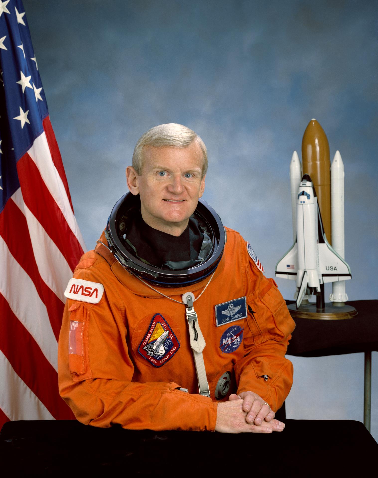 A man sits in front of the American flag and smiles, sitting next to a rocket ship model