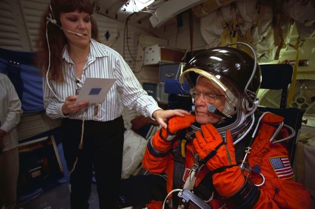 S98-06939 (28 April 1998) --- U.S. Sen. John H. Glenn Jr. (D.-Ohio) prepares to rehearse launch readiness procedures on the middeck of a crew trainer at the Johnson Space Center (JSC). Sharon Jones, involved in crew training, goes over a check list. When the STS-95 payload specialist lifts off aboard the Space Shuttle Discovery in October of this year and later lands in Florida, he will be seated in a temporary middeck chair like the one used in this training exercise. The photo was taken by Joe McNally, National Geographic, for NASA.