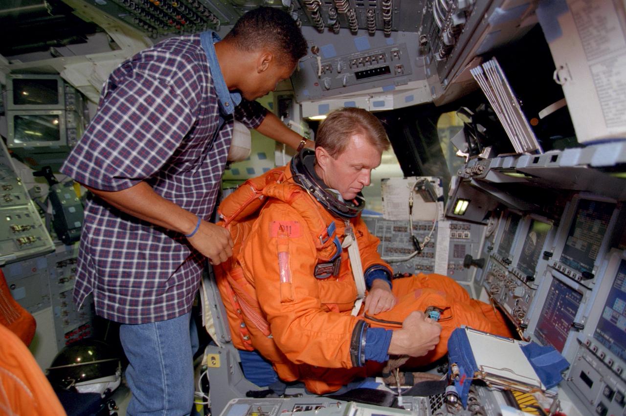 S99-07641 (15 July 1999)--- Carlous Gillis, a technician for United Space Alliance, assists astronaut Brent W. Jett, STS-97 commander, with his launch and entry garment during a training session in the Systems Integration Facility at the Johnson Space Center (JSC). Jett, pictured here in the commander's station on the port side of a shuttle crew compartment trainer (CCT), and four other astronauts are to visit the International Space Station (ISS) late next year