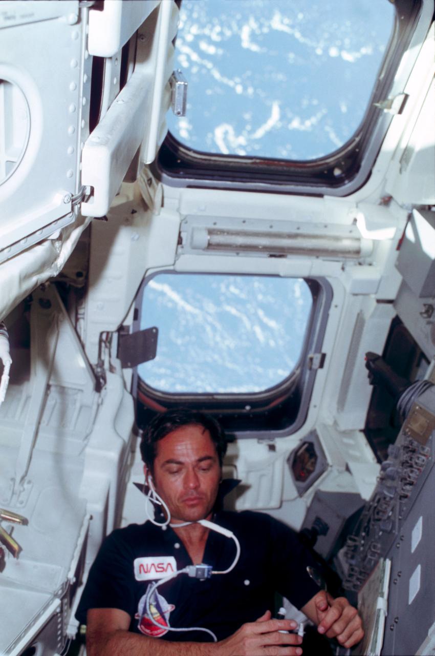 STS001-06-481 (April 1981) --- Astronaut Robert L. Crippen, STS-1 pilot, is behind his station in the flight deck of Columbia. He has a spoon in his hand. The windows prominent in the scene are the ones through which a number of Earth scenes were photographed with a 70mm camera by the two astronauts. Clouds over water can be seen through them. Just inches away from the top windows, not quite so obvious at top of the frame (if held horizontally) are the two aft cabin windows through which the crew viewed the cargo bay and the aft end of the craft, including the orbital maneuvering system (OMS) pods. Astronaut John W. Young, commander, took this photo with a 35mm camera. Photo credit: NASA