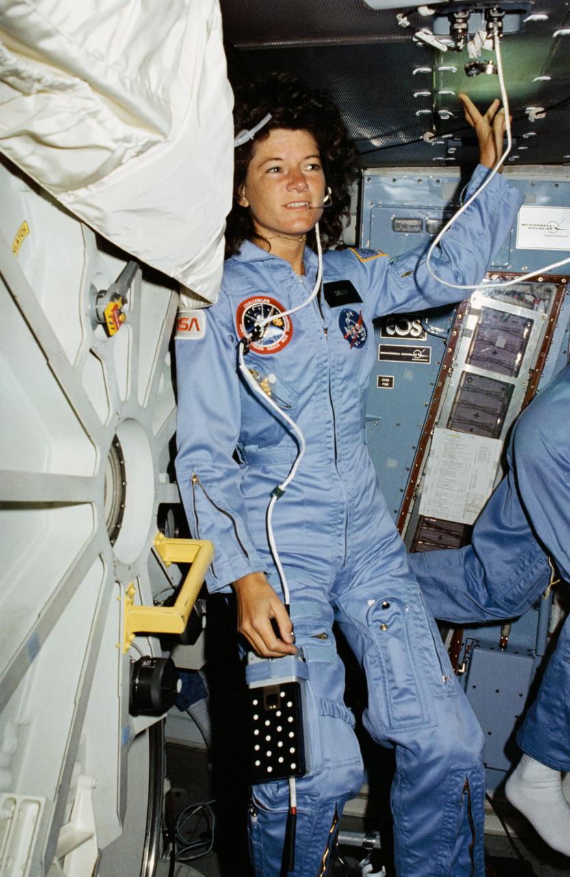 STS007-02-020 (21 June 1983) --- Astronaut Sally K. Ride, STS-7 mission specialist, STS-7 mission specialist, stands in the mid deck of the orbiting Space Shuttle Challenger near one of the experiment with which she has devoted a great deal of time.  The continuous flow electrophoresis system (CFES) experiment, about the size of a household refrigerator, stands nearby.  One of her fellow crewmembers moves partially out of frame in the background.  The tube on her face is part of a communications system linking Dr. Ride to ground controllers in Houston.