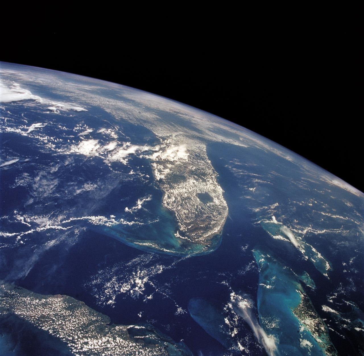 This unique photo offers a view of the Florida peninsula, western Bahamas, north central Cuba and the deep blue waters of the Gulf Stream, that hugs the east coast of Florida (27.0N, 82.0W). In addition to being an excellent photograph for showing the geographical relationships between the variety of landforms in this scene, the typical effect of the land-sea breeze is very much in evidence as few clouds over water, cumulus build up over landmass.