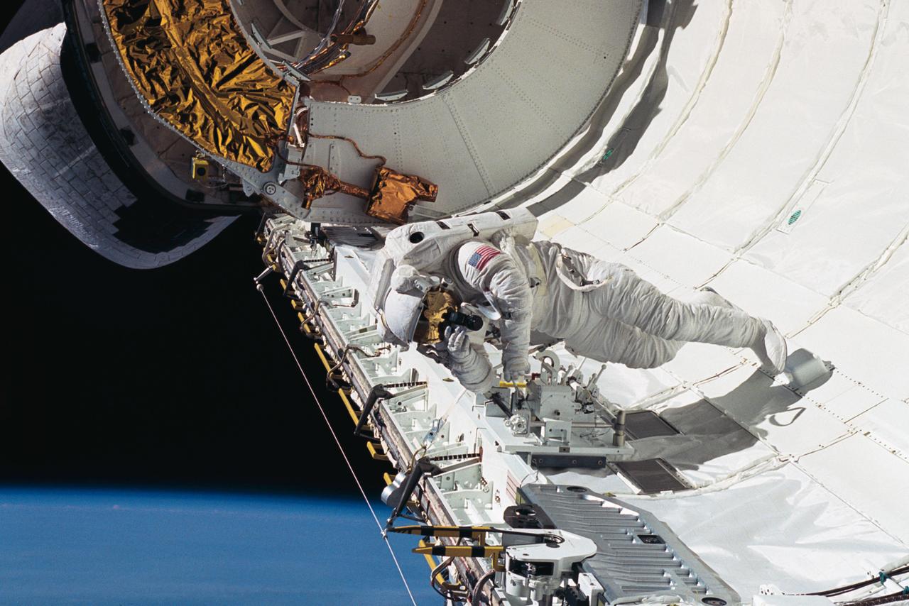 Astronaut James H. Newman, mission specialist, uses a 35mm camera to take a picture of fellow astronaut Carl E. Walz (out of frame) in Discovery's cargo bay. The two were engaged in an extravehicular activity (EVA) to test equipment to be used on future EVA's. Newman is tethered to the starboard side, with the orbital maneuvering system (OMS) pod just behind him.