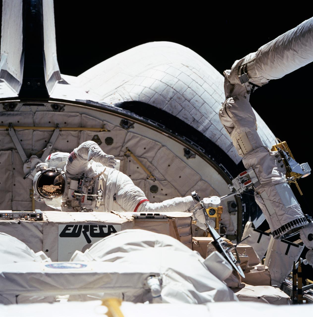The darkness of space forms the backdrop for this extravehicular activity (EVA) scene captured by one of the STS-57 crewmembers in Endeavour's, Orbiter Vehicle (OV) 105's, crew cabin. Pictured near the recently "captured" European Retrievable Carrier (EURECA) at frame center is Mission Specialist (MS) and Payload Commander (PLC) G. David Low. Suited in an extravehicular mobility unit (EMU), Low, anchored to the remote manipulator system (RMS) via a portable foot restraint (PFR) (manipulator foot restraint (MFR)), is conducting Detailed Test Objective (DTO) 1210 procedures. Specifically, this activity will assist in refining several procedures being developed to service the Hubble Space Telescope (HST) on mission STS-61 in December 1993. The PFR is attached to the RMS end effector via a PFR attachment device (PAD). Partially visible in the foreground is the Superfluid Helium Onorbit Transfer (SHOOT) payload.