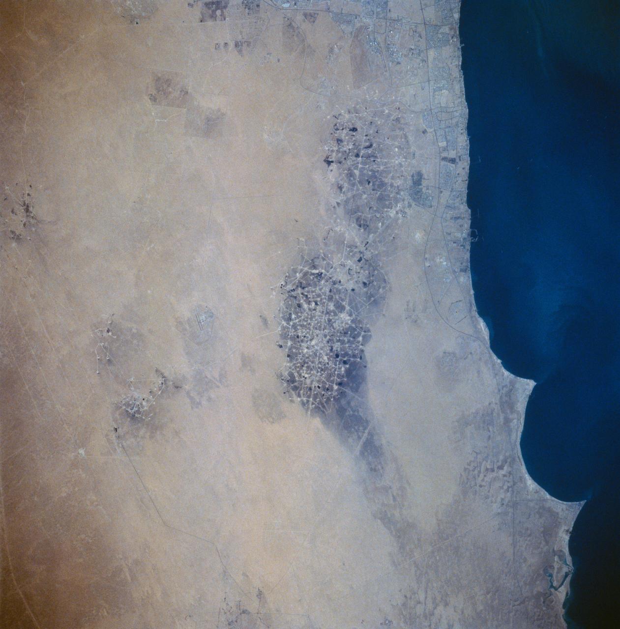 STS058-73-054 (18 Oct-1 Nov 1993) --- A clear view of the northern Kuwaiti coast shows the southern part of Kuwait City, and the major oil fields to the south.  Oil-laden sands, where wells were set ablaze during the Gulf War in 1991, are visible south of Kuwait City as a dark, elongated patch surrounded by light-colored sand.  Oil-stained sand between well sites (dots) and criss-crossing roads is gradually being covered by clean sand carried by strong, seasonal northwest winds.