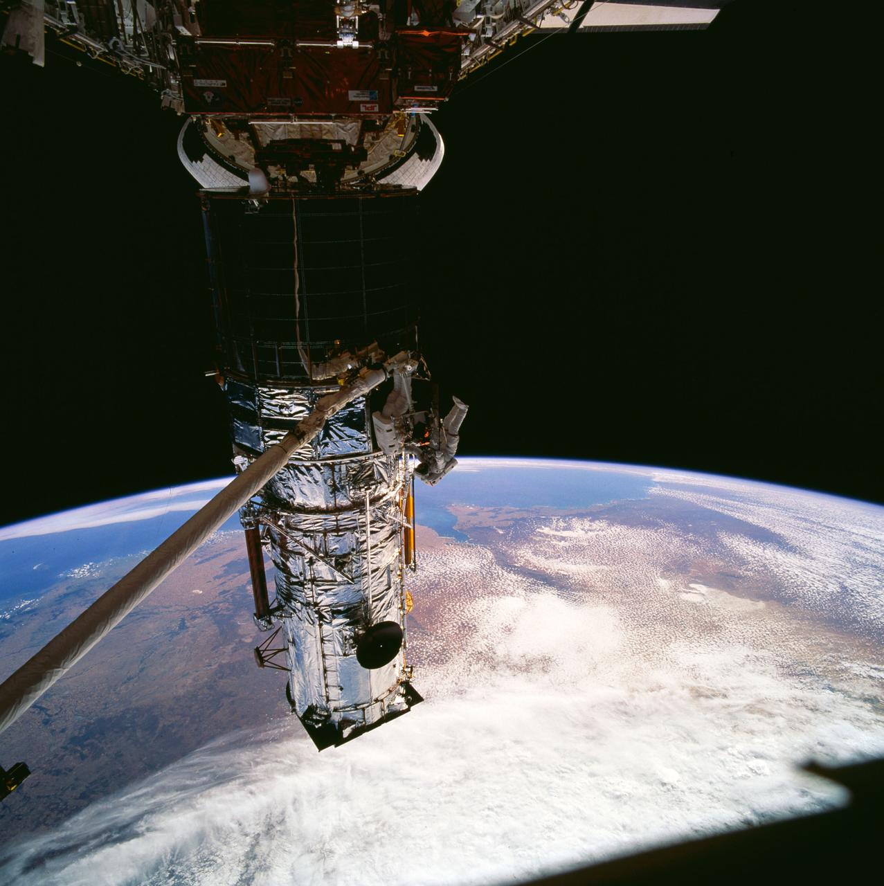 STS061-102-010 (9 Dec 1993) --- Astronauts Jeffrey A. Hoffman (left) and F. Story Musgrave team to replace one of two Solar Array Drive Electronics (SADE) units on the Hubble Space Telescope (HST).  Musgrave is standing on a foot restraint mounted on the end of the Space Shuttle Endeavour's Remote Manipulator System (RMS) arm.  The black object, in upper left corner, is part of the window frame, through which this 70mm frame was exposed, inside Endeavour's cabin.
