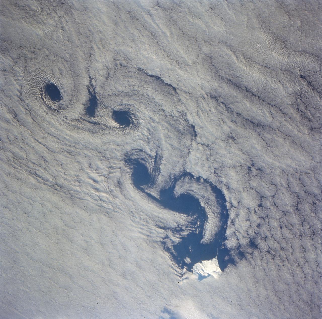 As the air blows over the tops of the mountain peaks, seen here in the clear region, the direction of the air flow is disturbed. These disturbances in the flow pattern are generally small circular eddies with one circulating in a clockwise direction and the next in the reverse direction. The wind here has caused the shallow cloud cover to change accordingly. These eddies are commonly called von Karmen Vortices.