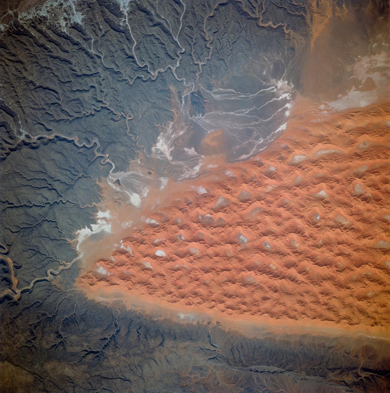 STS070-705-094 (13-22 JULY 1995) --- The southern half (about 70 miles in this view) of the Tifernine dunes of east-central Algeria appears on this view. The Tifernine dune-sea is one of the more dramatic features visible from the Shuttle when flying over the Sahara Desert. The dunes lie in a basin of dark-colored rocks heavily cut by winding stream courses (top right). Very occasional storms allow the streams to erode the dark rocks and transport the sediment to the basin. Westerly winds then mold the stream sediments into the complex dune shapes so well displayed here. North at bottom.