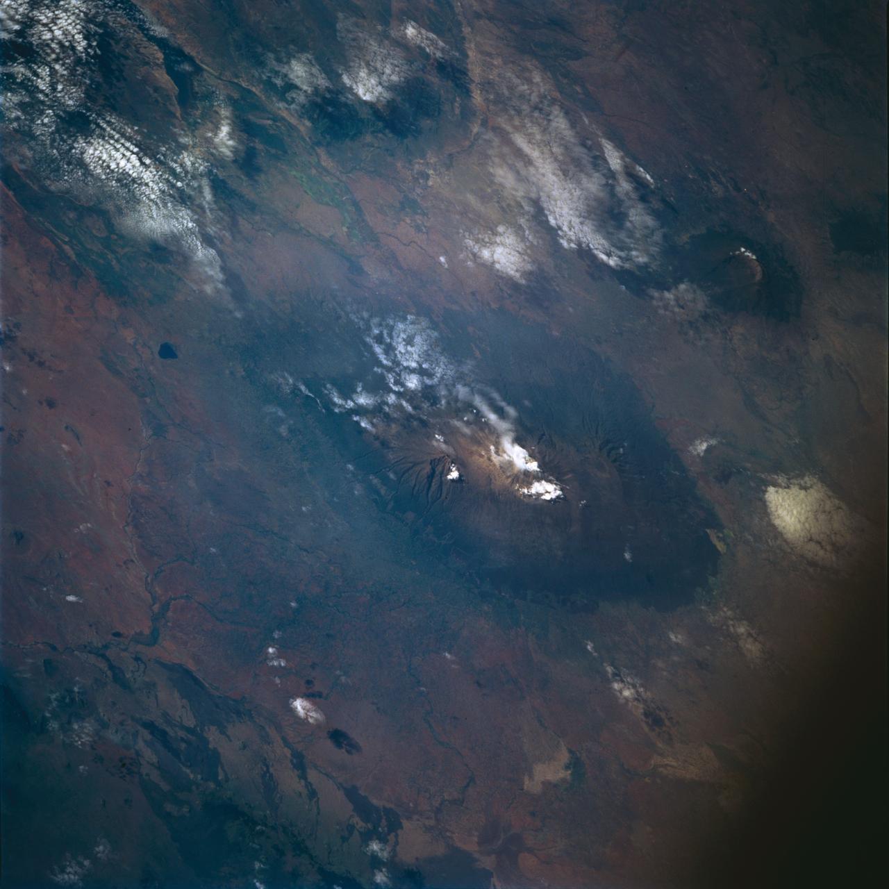 STS072-722-004 (11-20 Jan. 1996) --- Mount Kilimanjaro in Tanzania is featured in this 70mm frame exposed from the Earth-orbiting Space Shuttle Endeavour.  Orient with the clouds trailing to the left; then the view is southwest from Kenya past Kilimanjaro to Mount Meru, in Tanzania.  Mount Kilimanjaro is about three degrees south of the Equator, but at nearly 6,000 meters has a permanent snowfield.  The mountain displays a classic zonation of vegetation types from seasonally dry savannah on the plains at 1,000 meters, to the cloud forest near the top.  The mountain is being managed experimentally as an international biosphere reserve.  A buffer zone of "traditional" agriculture and pastoral land use is designated around the closed-canopy forest reserve.  Specialists familiar with this area say management is partially successful so far, but cleared areas of the forest can be seen on this photograph as light green "nibbles" or "cookie cuts" extending into the dark forest region.