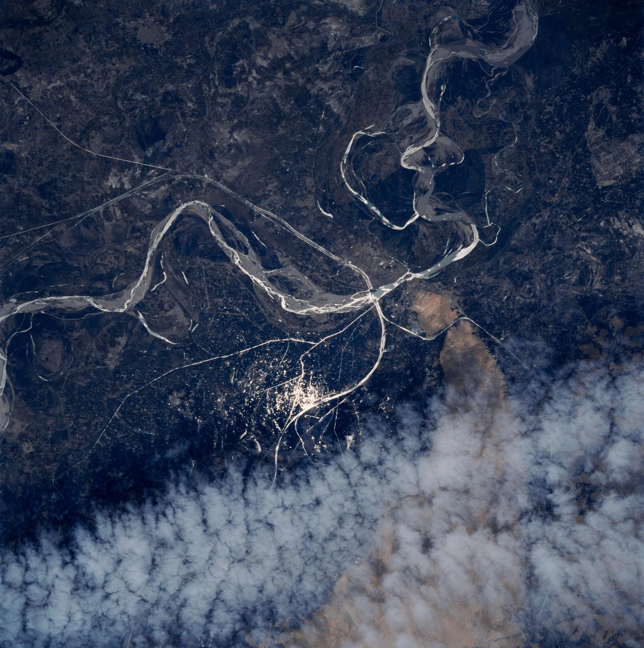 STS083-747-052 (4-8 April 1997) --- Sunglint on the Indus River, Sukkar, and Rohri, Pakistan.  Sukkar city (27.42 north 68.52 east), Sindh province, southeastern Pakistan lies on the west bank of the Indus River, connected with Rohri on the opposite bank by a cantilever bridge.  Midstream between the two cities is the strategic island fortress of Bukkur.  The old town contains many historic tombs and mosques, including the Mir Ma'sum Shah Minaret (c. AD 1607).  An industrial and trade center, it has biscuit, cigarette, oil, lime, and cement factories, and cotton, silk, thread, and flour mills; boat building is also significant.  The surrounding region is a vast alluvial plain broken only occasionally by low limestone hills.  A portion of the Thar Desert is reaching from the south to Rohri.  The Sukkur Barrage, highlighted by the sunglint, was completed in 1932.  Nearly 1 mile (1.6 kilometers) long it crosses the Indus River 3 miles (4.8 kilometers) below Sukkur Gorge and feeds irrigation canals.  The canals originating from it serve a cultivable area of about five million acres of land producing both food and cash crops, such as wheat, cotton, rice, oilseed, and fruit cultivation.
