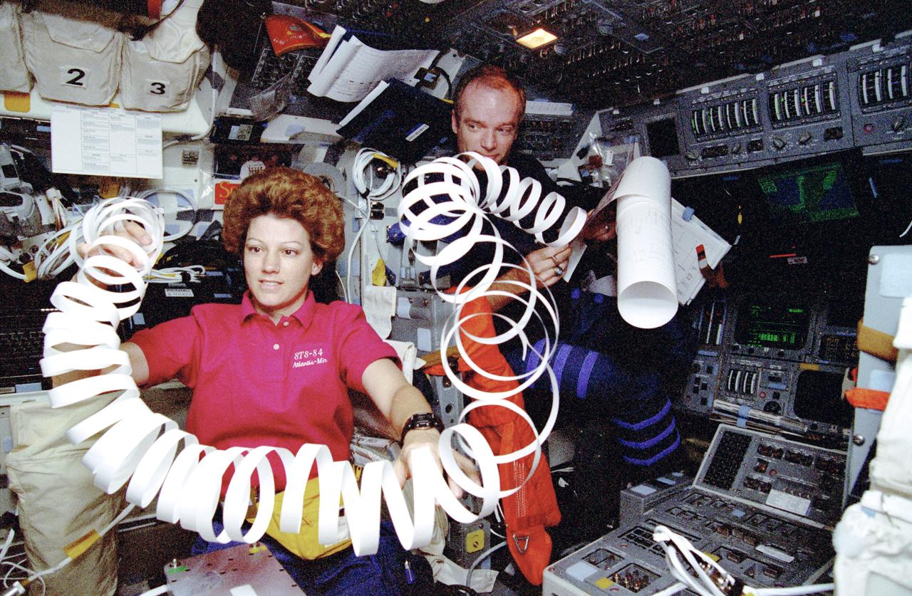 STS084-358-009 (15-24 May 1997) --- Astronaut Eileen M. Collins, STS-84 pilot, handles what appears to be an oversized space toy but is actually a lengthy roll of paper margin, separated from the main roll of messages just transmitted to the flight crew from ground controllers in Houston, Texas.  Seated at the commander's station, astronaut Charles J. Precourt, commander, checks the latest transmission from a message device called Thermal Imaging Printing System (TIPS).
