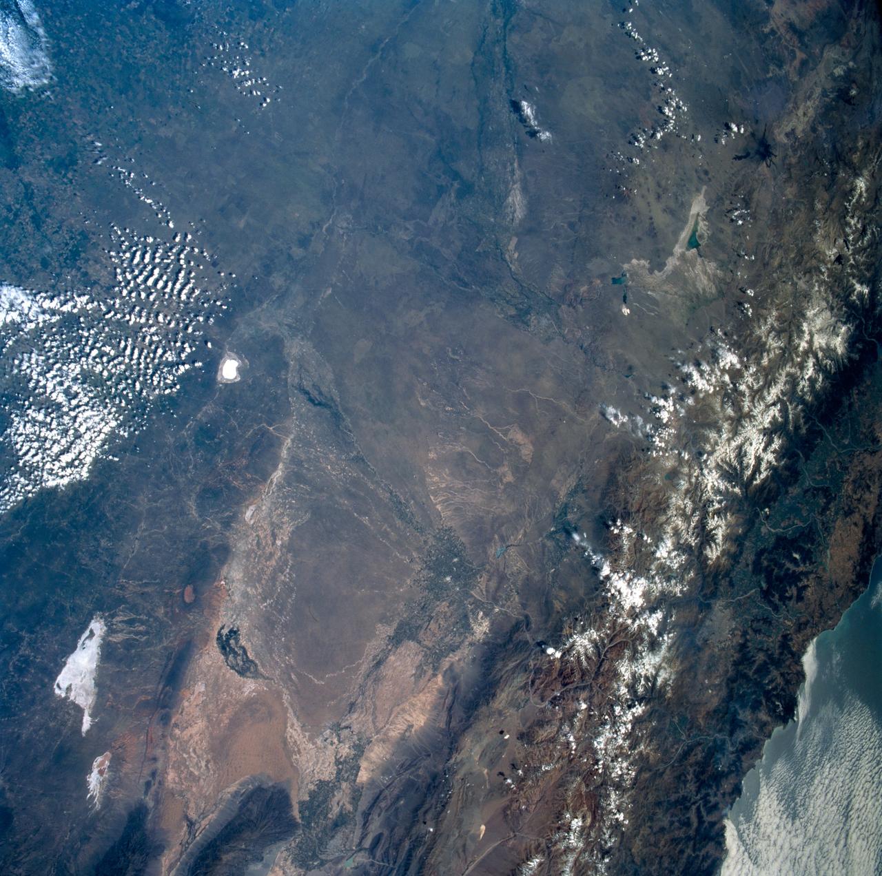 STS089-742-014 (22-31 Jan. 1998) --- This near-nadir picture of the central Andes, showing Santiago and other parts of Chile and Argentina, was photographed with a 70mm handheld camera from the Earth-orbiting space shuttle Endeavour. Santiago (population 4.7 million) is visible to the right of center, west of the snow-capped Andes Mountains. Mendoza, Argentina is the light spot in the upper center of the image in the eastern foothills of the Andes. Cerro Aconcagua, the highest peak in South America (22,834 feet) is due west of Mendoza. Note the sharp bend in the river valley to the junction with a straight north-trending valley. Aconcagua is the large, rounded mass with a bit of cloud just northwest of the valley junction.  Bright white spots on the left of the image are salars, or dry lake beds of gypsum and/or salt.  Usually dry, some contain water due to recent rains generated by the record El Nino this year. The Pacific Ocean is to the upper right.  The continental shelf is very steep in this region and water depths drop (to almost 17,000 feet over a distance of about 60 miles) over the same distance inland from the coast, the Andes rise +17,000 to almost 23,000 feet. The reason for this is the very rapid plate motion between the Pacific Ocean plate and South America.  According to NASA scientists, over the last 140 million years, South America has been overriding the Pacific plate resulting in the rapid rise in the Andes and the rapid subduction of the Pacific plate, along with any sediments that are shed into the ocean along the continental margin.  The dark, linear mountain ranges east of the high Andes (right center) constitute the Precordillera block; rock strata there have been correlated with the Ordovician (approximately 470 million years old) rocks near El Paso, Texas. The rocks and the fossils within them are virtually identical in the two areas, which are now widely separated. This region would be considered a "remnant terrain" from the time when this portion of South America and Texas were once joined.  Photo credit: NASA