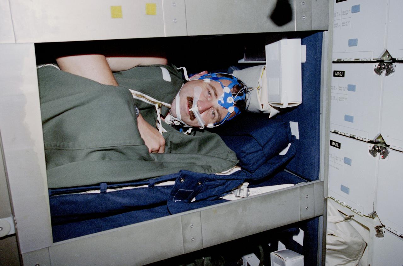 STS090-377-011 (17 APRIL-3 MAY 1998) --- Astronaut Dafydd R. (Dave) Williams, mission specialist representing the Canadian Space Agency (CSA), accomplishes more than one purpose when he sleeps in this bunk aboard the Earth-orbiting Space Shuttle Columbia.  Conducting a Neurolab sleep experiment, Williams wears equipment which includes a sleep net (mesh cap that monitors and records brain waves); a Respiratory Inductance Plethysmograph (RIP) suit for monitoring respiration; and an activity monitor -- a device (out of view) worn on the wrist to detect and record body movement.  Data on brain waves, eye movements, respiration, heart rate, and oxygen concentration are routed to a portable data recorder.  The entire system has capabilities similar to a fully equipped sleep laboratory on Earth.  The sleeping bag is conventional Shuttle ware and not part of the experiment.