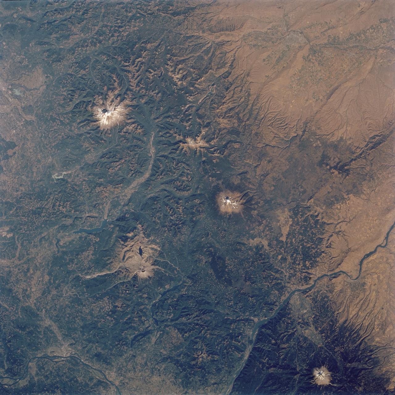 STS112-704-142  (7-18 October 2002)  ---  (For orientation purposes, north is toward the top left corner).  Green colors of the forests of the Cascade Mountains dominate this view, photographed from the Earth-orbiting Space Shuttle Atlantis.  Browner colors (top right) are the semiarid plains of the Columbia Basin, in the rain shadow of the Cascades. The highest peaks in this part of the Cascades are four volcanoes.  The amount of snow is a good indication of their altitude.  The highest is Mt. Rainier (14,410 feet) with the greatest amount of white snow (top left).  Seattle lies immediately downslope (top left margin).  Mt. Adams (12,276) lies due south in the middle of the view.  Mt. Hood (11,235 feet) in the lower right corner, lies south of the great gorge of the Columbia River (which crosses the lower right and then the lower left corners of the view). The river flows broadly west (left) to the Pacific Ocean (out of the picture left).  Mt. St Helens (8,364 feet), the snow-free brown patch lower left, was too low to retain snow after the recent fall.  According to geologists studying the STS-112 photography, even from the altitude of the Space Shuttle, the intact south half of the cone can be discerned.  The geologists point out that  the famous blast of 1980 not only destroyed the north side of the cone but blew down the green forest for many square miles on the north side (brown signature).