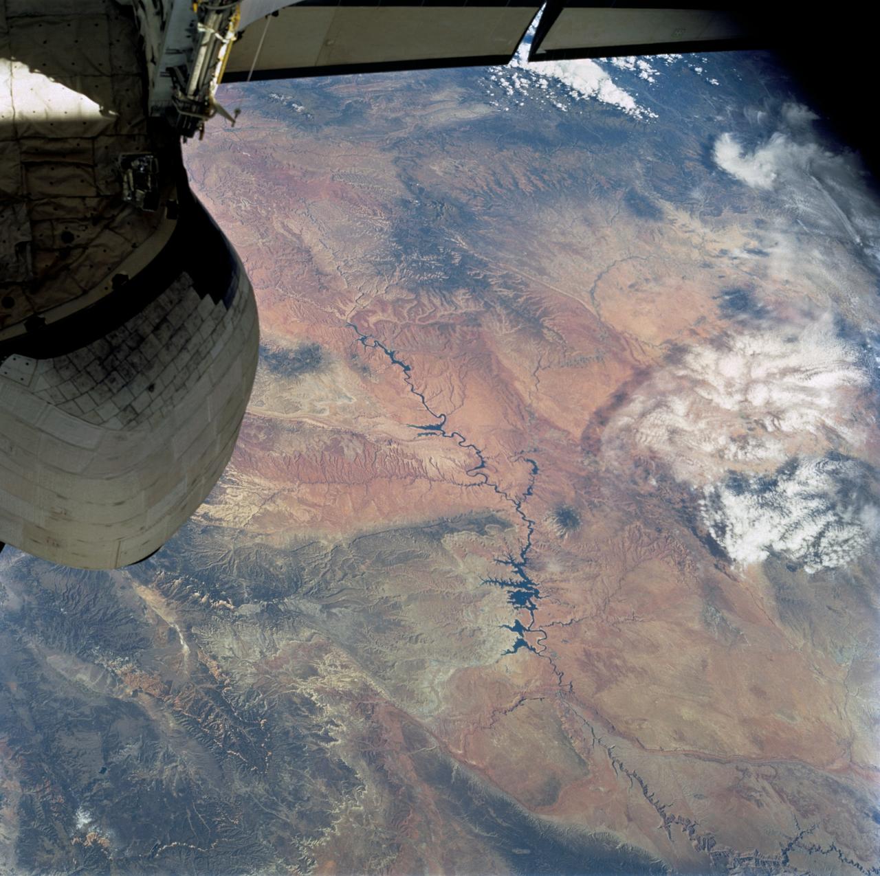 STS112-707-035 (7-18 October 2002) --- The Colorado River snakes across this view, photographed from the Space Shuttle Atlantis, from top left (near the vehicle’s vertical stabilizer), to the lower right, where the Grand Canyon gorge can be detected. The wider blue parts of the Colorado in the center of the view are Lake Powell, an artificial lake more than 120 miles long. The dark green areas are wooded slopes generally above 6000 feet. The Kaibab Plateau (lower left) and the range with Abajo Peak (11,360 feet, Utah-Colorado border region) cross the top of the view. The red areas are lower and semiarid, dominated by desert scrub vegetation.